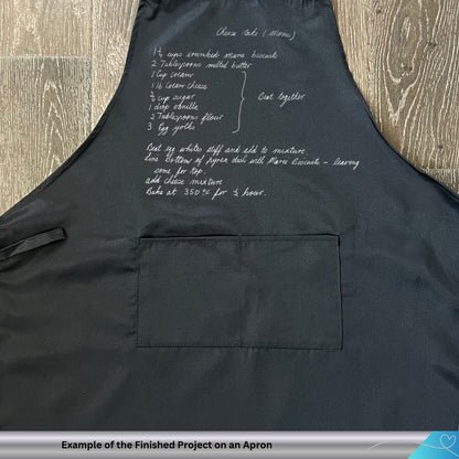 Black apron with white text hand embroidered on a wooden surface