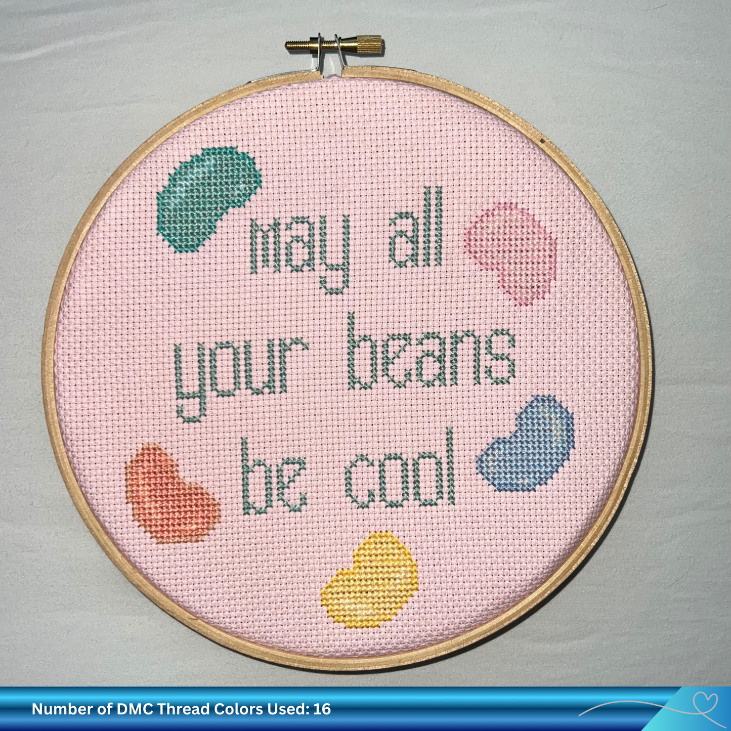 Cross-stitched hoop with 'May all your beans be cool' text on a pink background.
