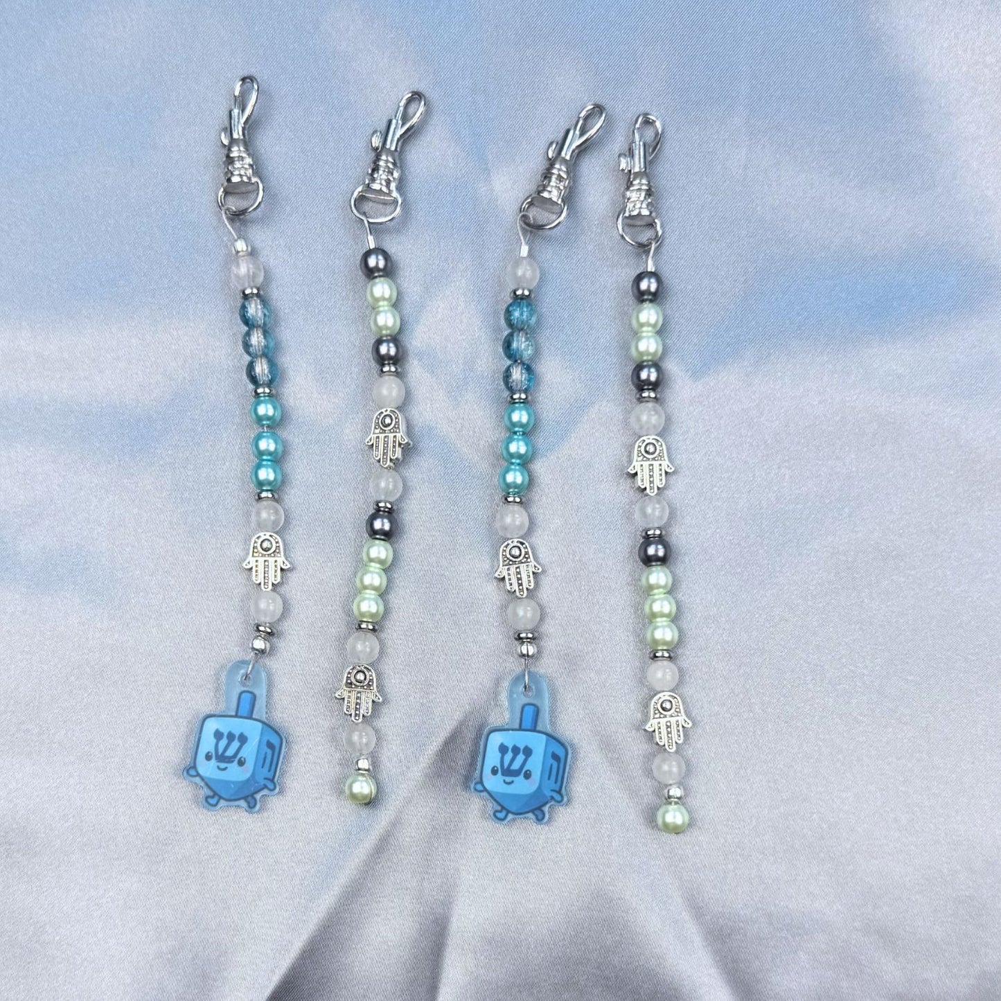 Four beaded keychains with blue dreidel and hamsa charms on a light blue background.