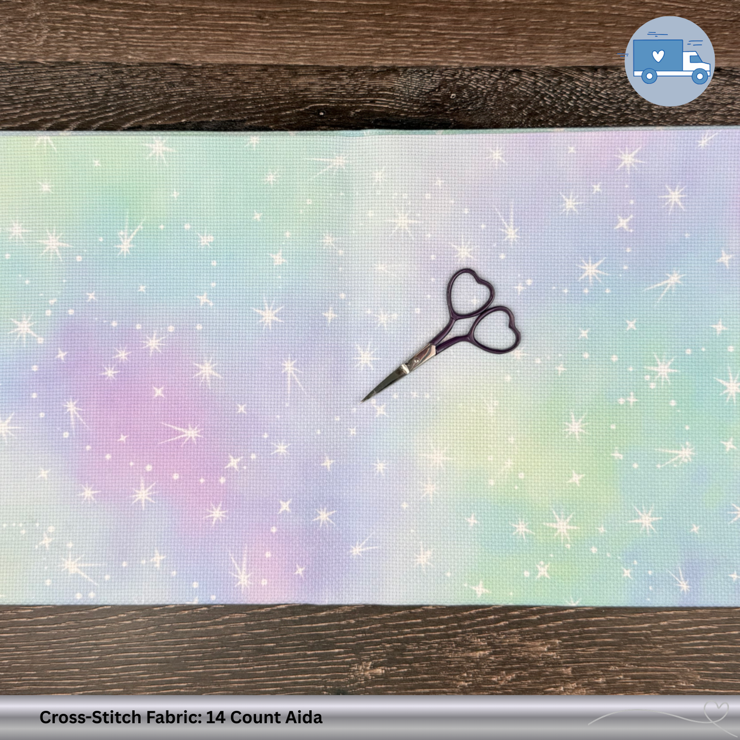 Colorful pink, green, blue, purple cross-stitch fabric with glitter and sparkles. A dark purple pair of scissors sits on top of the fabric on a wooden surface