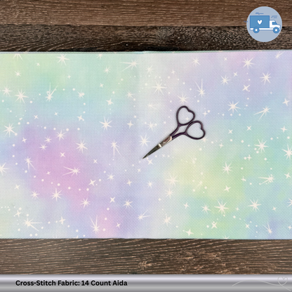 Colorful pink, green, blue, purple cross-stitch fabric with glitter and sparkles. A dark purple pair of scissors sits on top of the fabric on a wooden surface