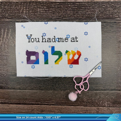 Cross-stitched funny text saying "You had me at Shalom" in rainbow colorful letters on a wooden surface with scissors.