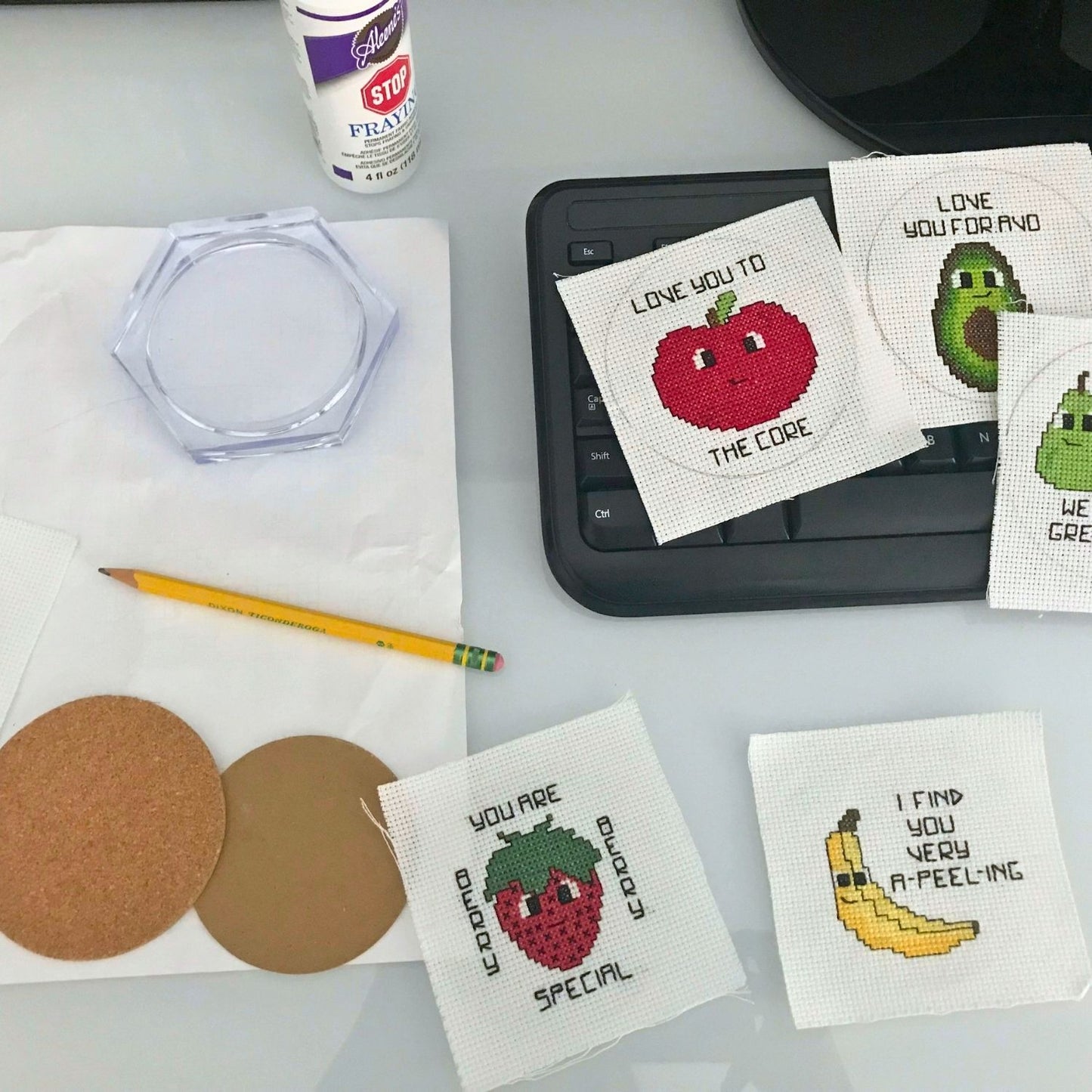 Cross-stitched coasters with fruit designs on a table with a laptop and stationery.