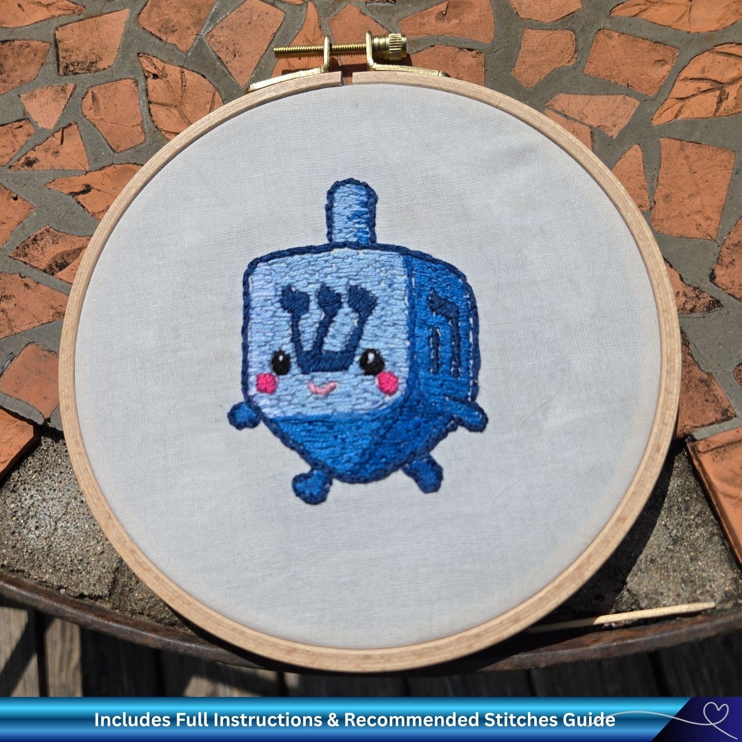 Embroidery hoop with a Chanukah dreidel design on fabric against a stone wall background.