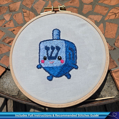 Embroidery hoop with a Chanukah dreidel design on fabric against a stone wall background.