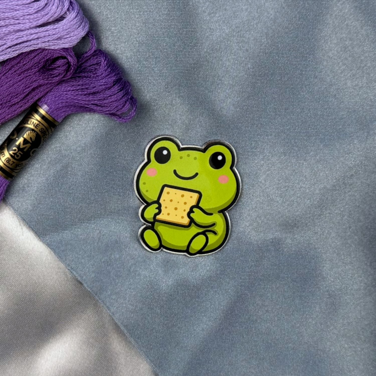 Green frog needle minder holding a piece of Matzah on a gray fabric background with purple and gold thread spool.