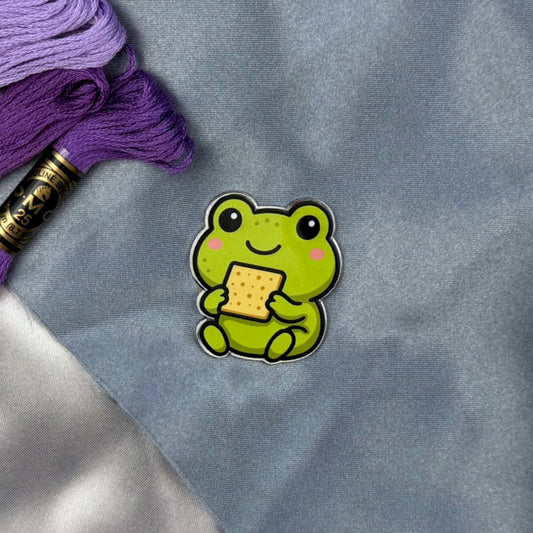 Green frog needle minder holding a piece of Matzah on a gray fabric background with purple and gold thread spool.