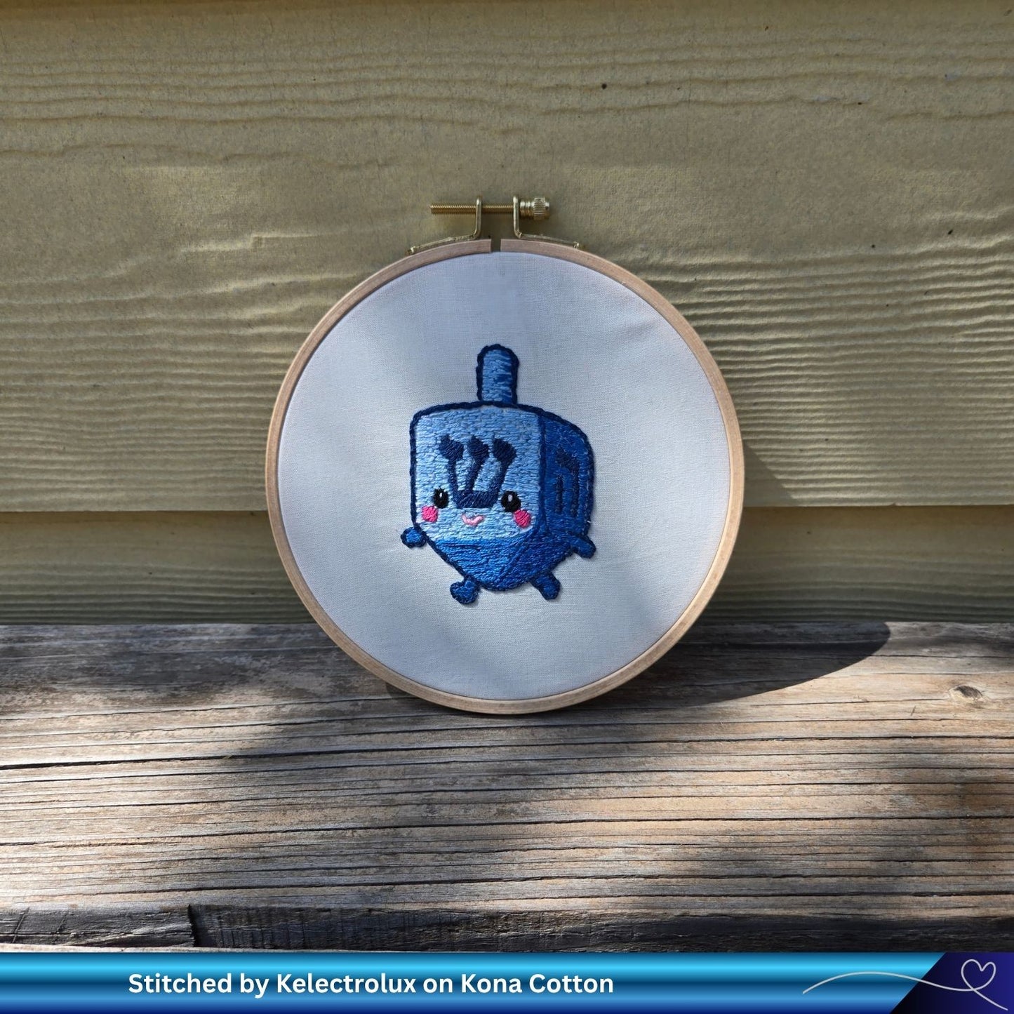 Embroidery hoop with a blue dreidel character design on a wooden surface