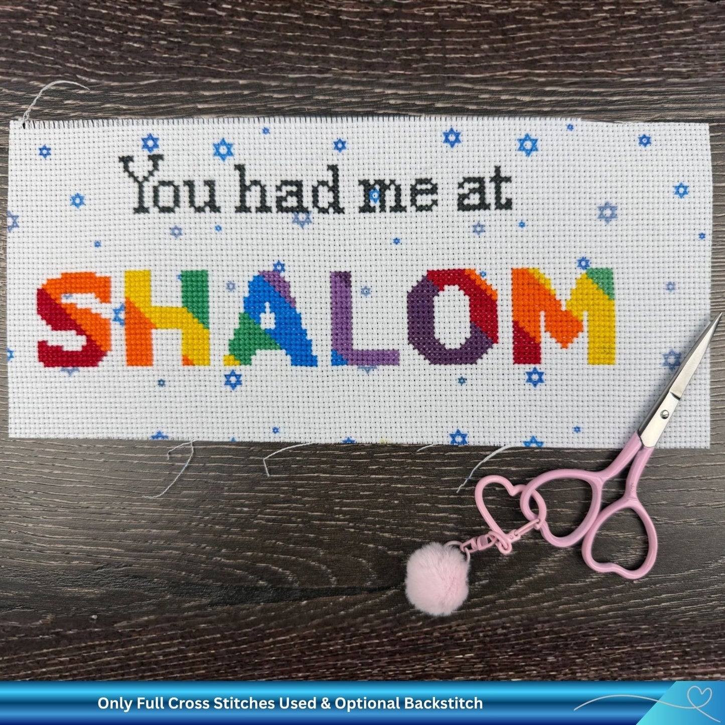 Cross-stitched Aida fabric with 'You had me at SHALOM' text on a wooden surface with scissors.