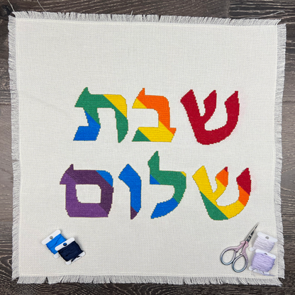 Colorful Hebrew text saying Shabbat Shalom on a white fabric with embroidery tools on a wooden surface