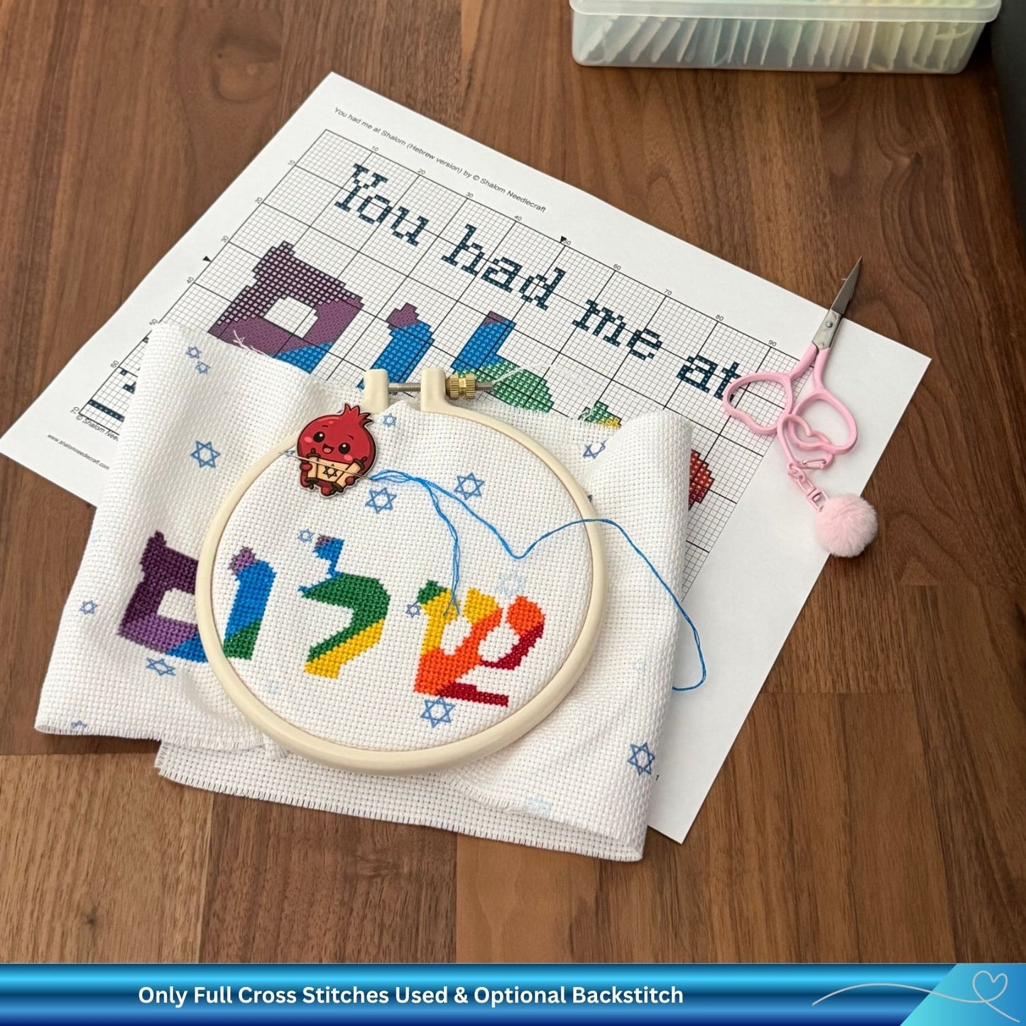 Embroidery hoop with colorful letters on a wooden table with cross-stitch pattern and embroidery scissors.