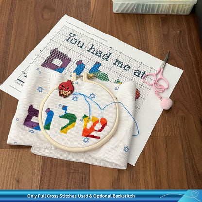 Embroidery hoop with colorful letters on a wooden table with cross-stitch pattern and embroidery scissors.
