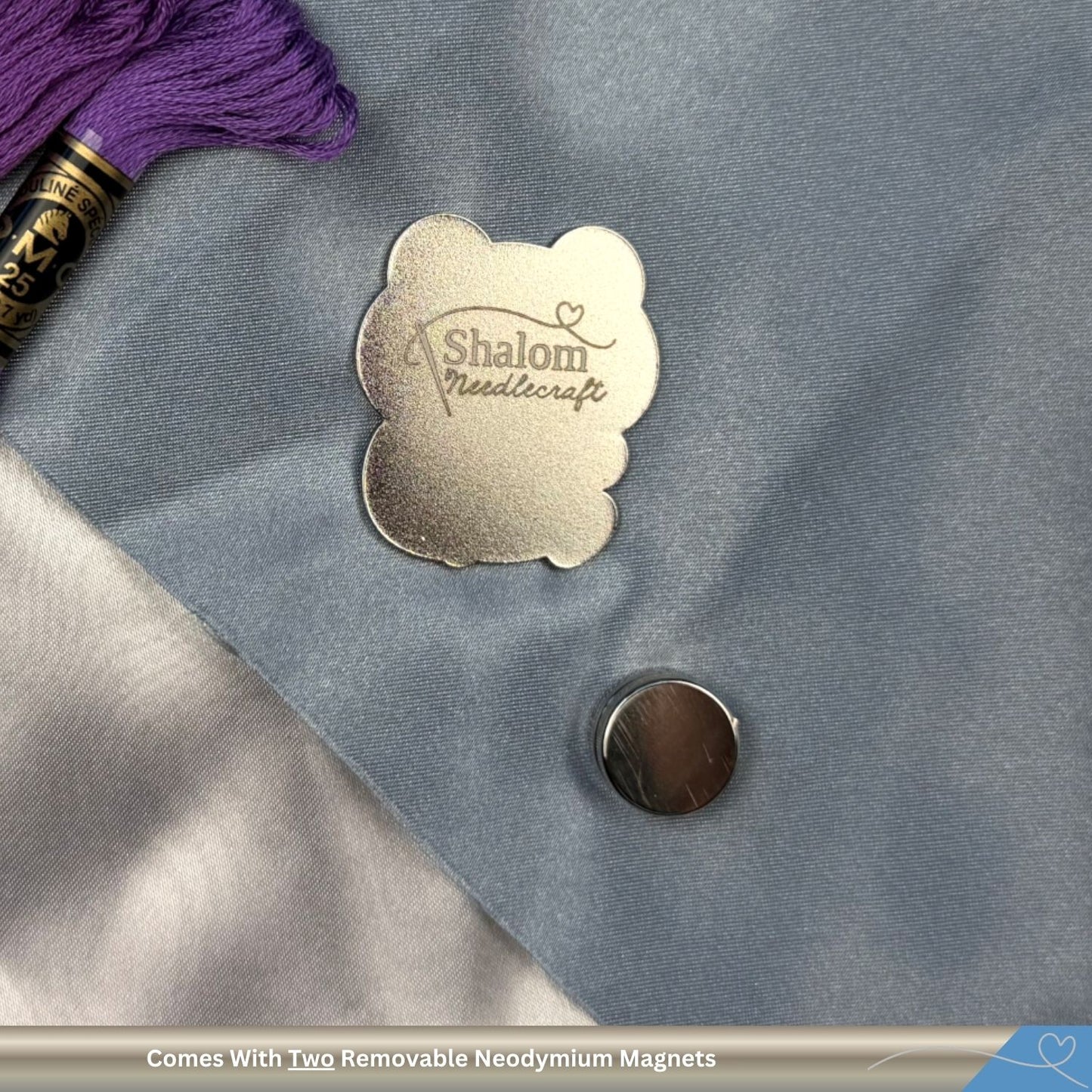 Metallic needle minder with 'Shalom Needlecraft' on a textured surface with purple thread and a magnet.