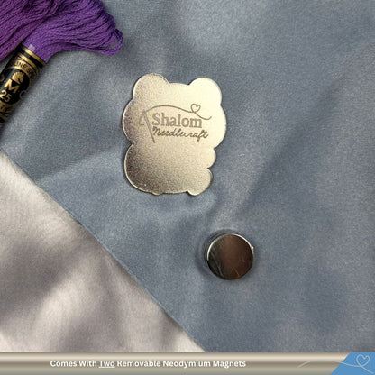 Metallic needle minder with 'Shalom Needlecraft' on a textured surface with purple thread and a magnet.