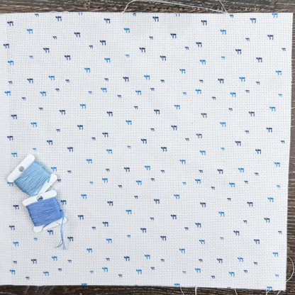 White cross stitch fabric with blue Chai pattern and two spools of blue thread on a wooden surface