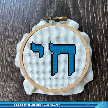 Cross-stitch hoop with blue Hebrew word for Life on white Aida fabric against a wooden background