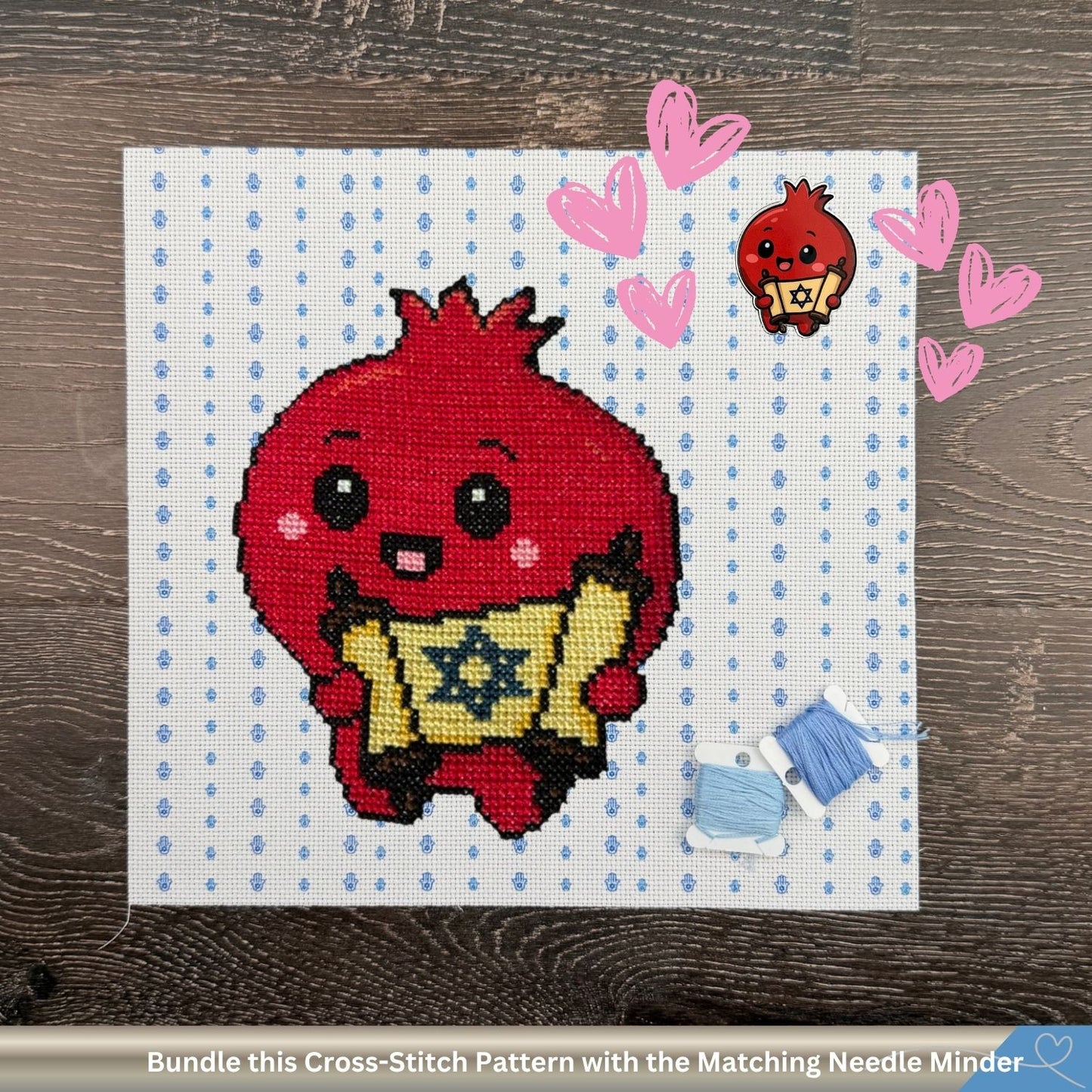 A cross-stitch pattern display featuring a cute red pomegranate character holding a yellow Star of David scroll, shown on a white and blue Hamsa cross-stitch canvas. The canvas is placed on a dark wood background, surrounded by pink hearts and small accessories including blue thread and a matching small red pomegranate needle minder.