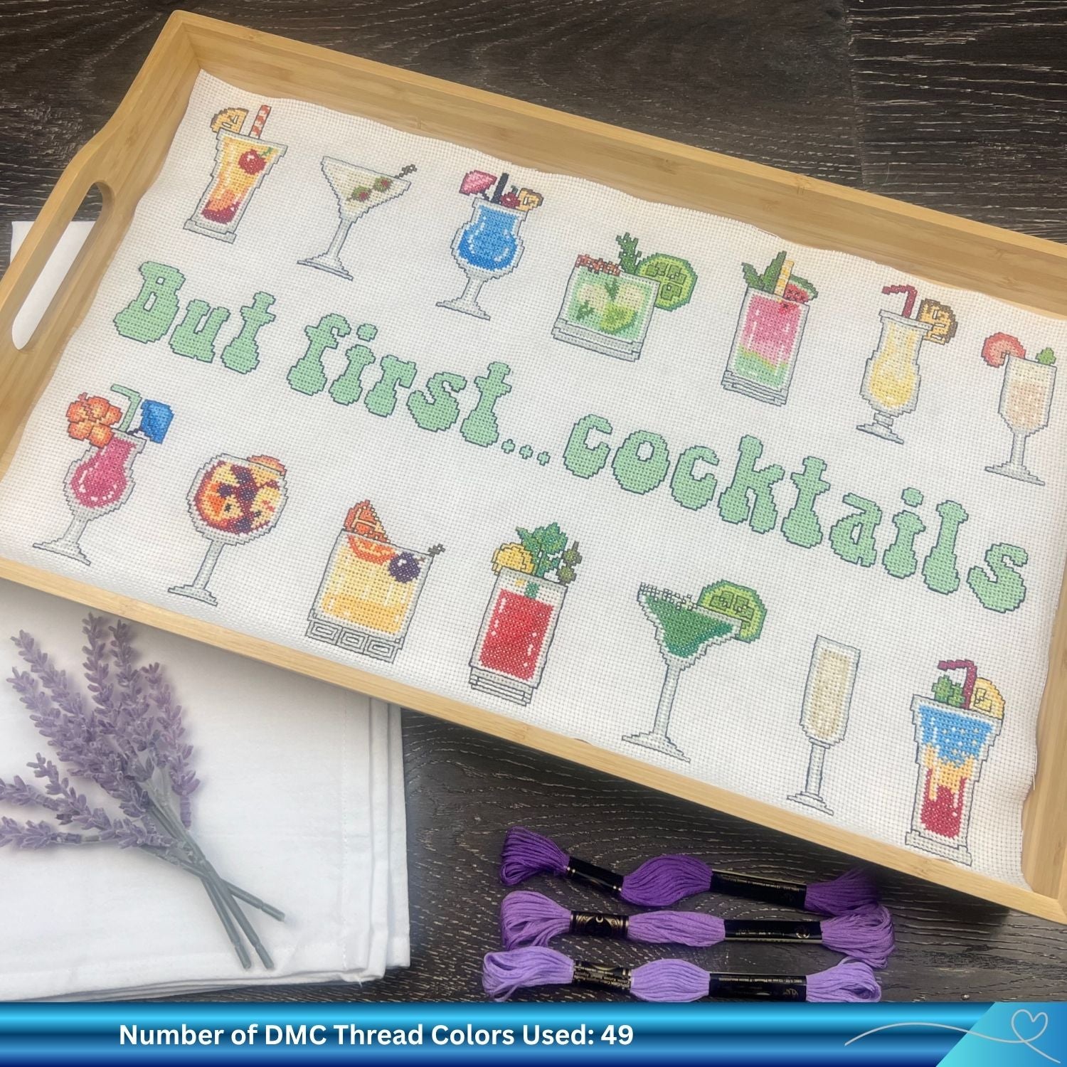 Cross-stitched tray with cocktail drink designs and text 'But first... cocktails' on a wooden tray. There is purple DMC thread towards the bottom of the image on a wooden floor.