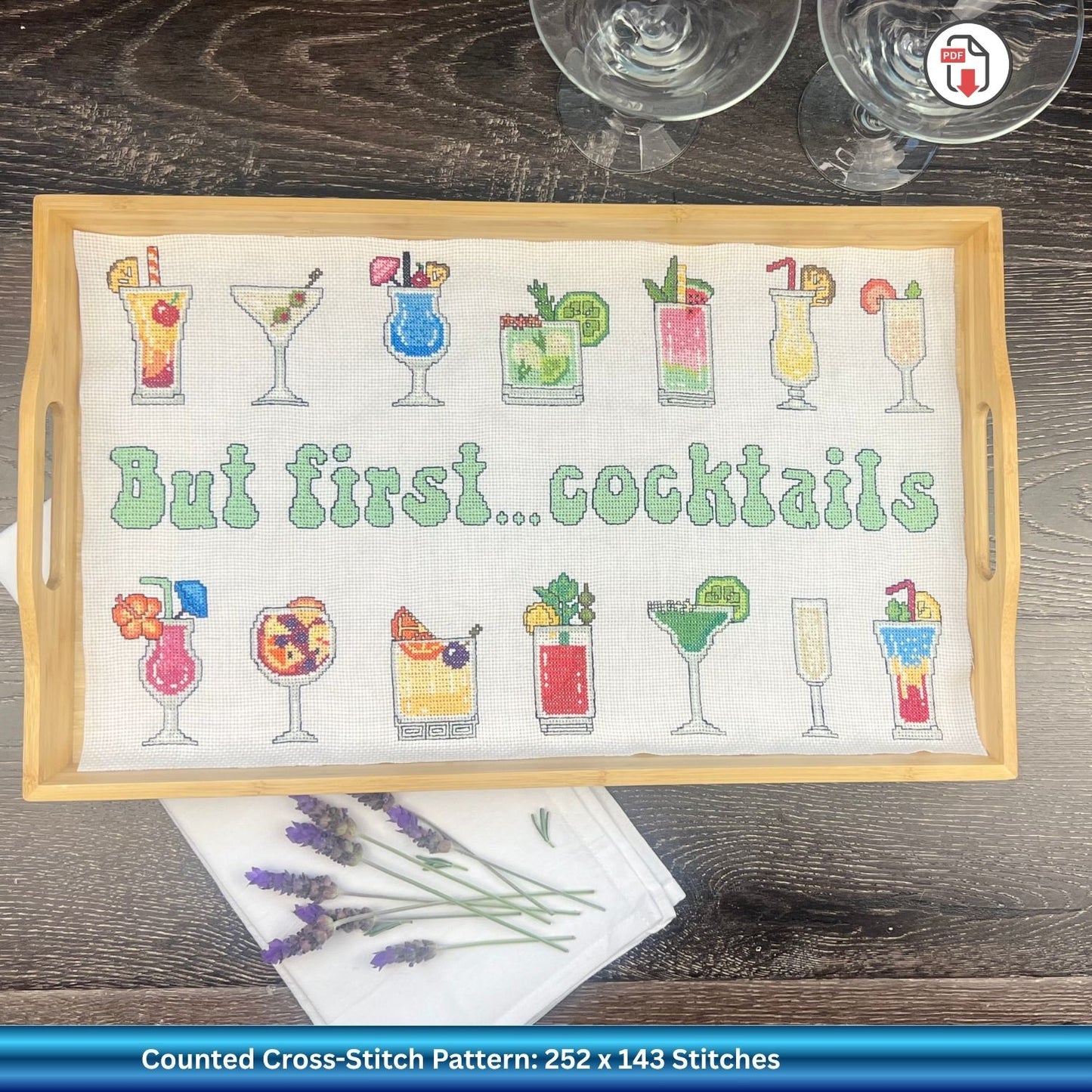 Cross-stitched tray with cocktail designs and 'But first... cocktails' text on a wooden tray against a wooden background. There are cocktail glasses at the top right and lavender flowers towards the bottom left.