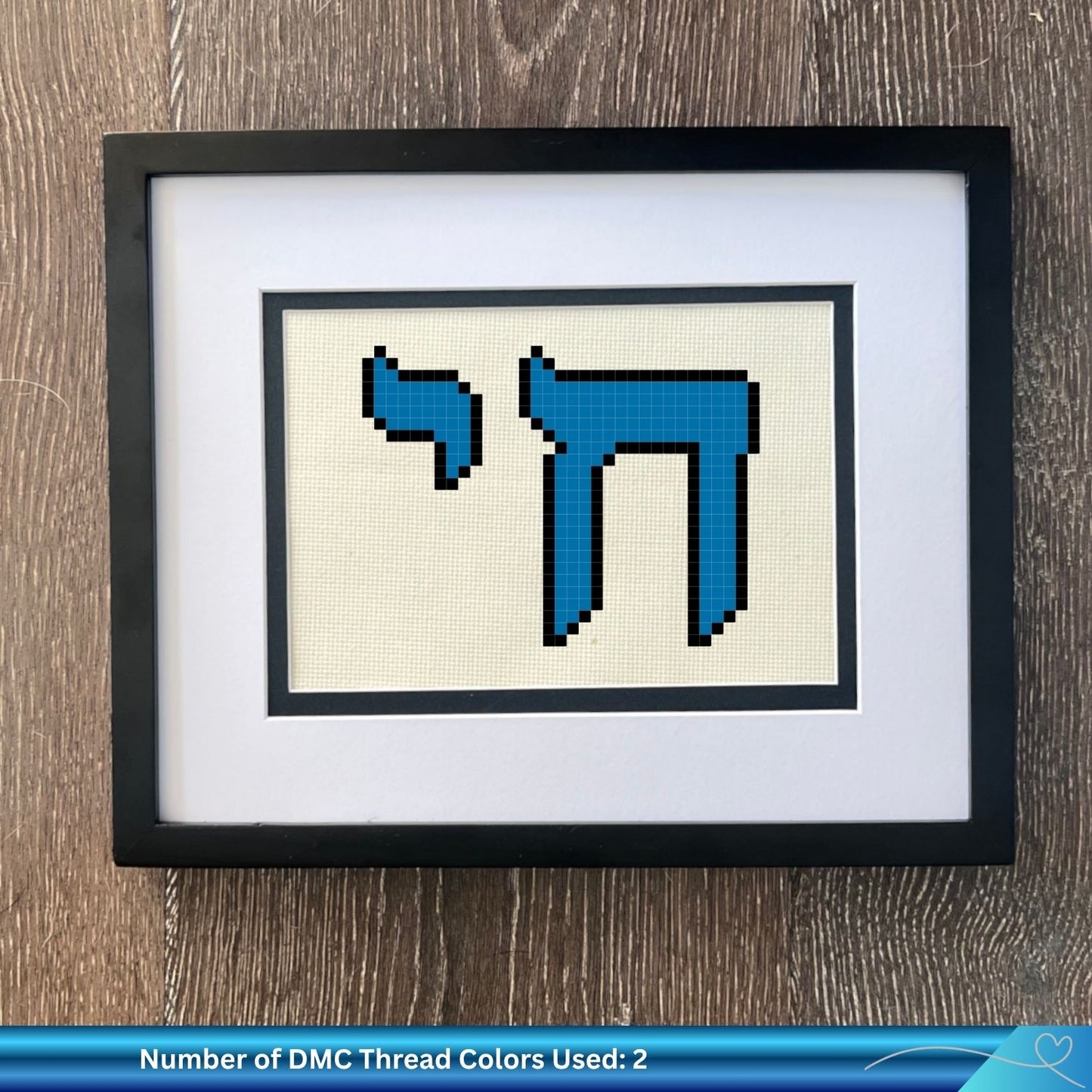 Framed cross-stitched design with the word Chai in Hebrew letters on beige fabric, placed on a wooden surface.