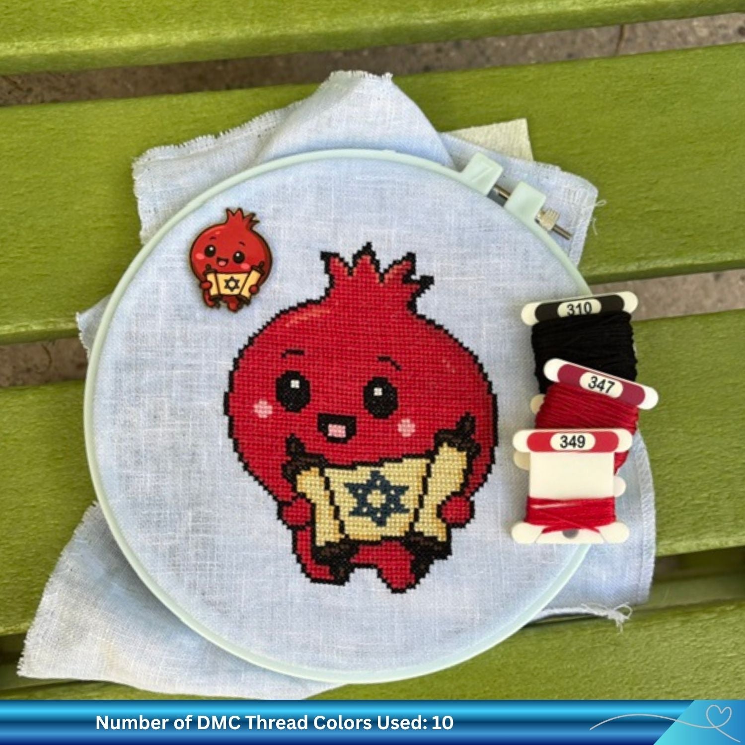 A circular embroidery hoop holding light blue linen fabric, featuring a stitched red cartoon pomegranate holding a golden scroll with a Star of David. A small red pomegranate needle minder is placed beside the hoop, and three spools of DMC thread in black and two shades of red are arranged on the right.