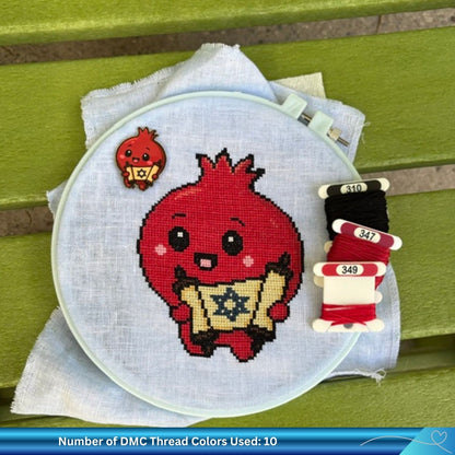 A circular embroidery hoop holding light blue linen fabric, featuring a stitched red cartoon pomegranate holding a golden scroll with a Star of David. A small red pomegranate needle minder is placed beside the hoop, and three spools of DMC thread in black and two shades of red are arranged on the right.