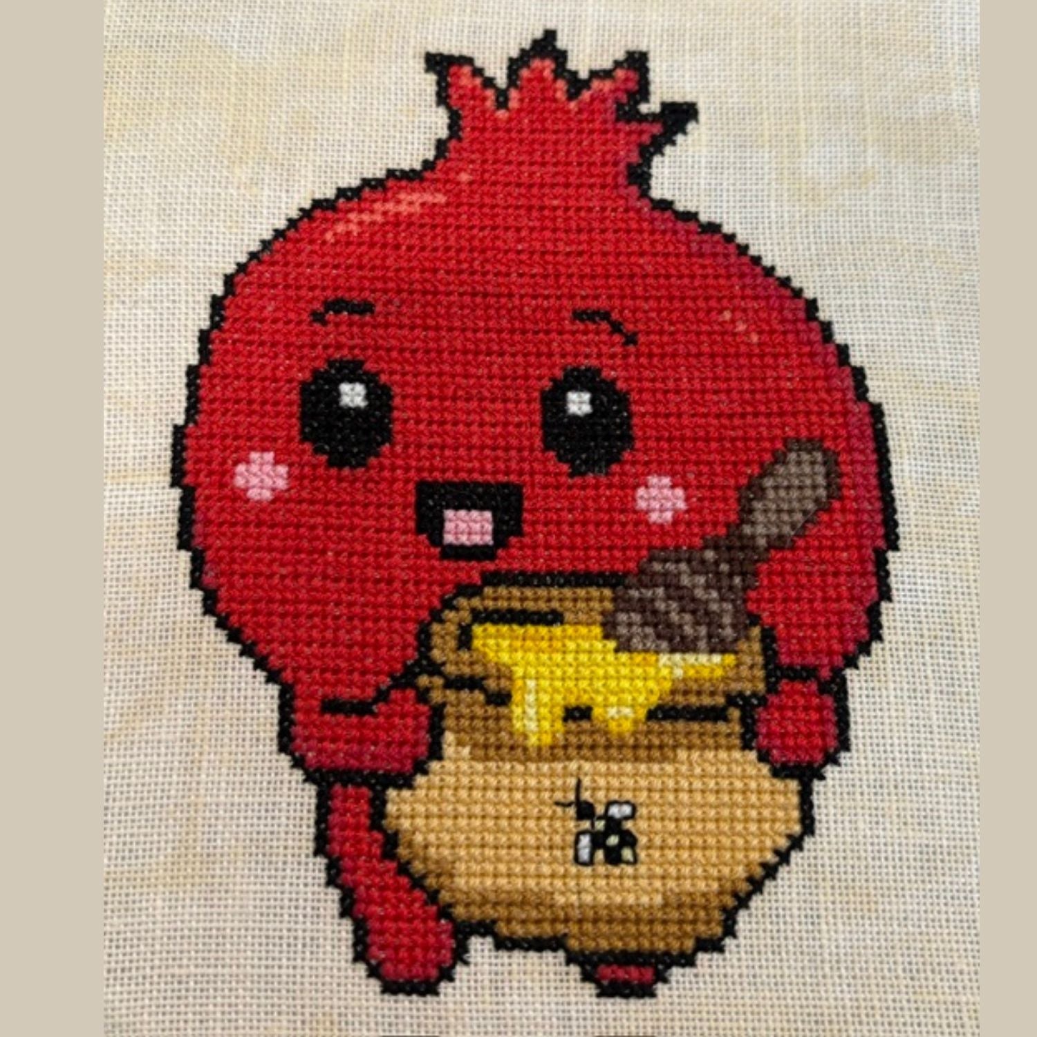 A cross-stitch or needlepoint artwork on yellow cream fabric showing a round red pomegranate character with black eyes, pink cheeks, and a smiling mouth, holding a honey pot and a wooden honey spoon. The piece is photographed close-up against a light beige fabric background.