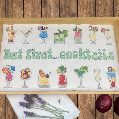 Cross-stitched design with cocktail glasses and 'But first... cocktails' text lining a wooden tray. There are passion fruit and lavender flowers around the tray against a wooden surface.
