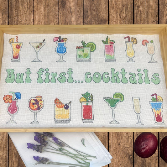 Cross-stitched design with cocktail glasses and 'But first... cocktails' text lining a wooden tray. There are passion fruit and lavender flowers around the tray against a wooden surface.