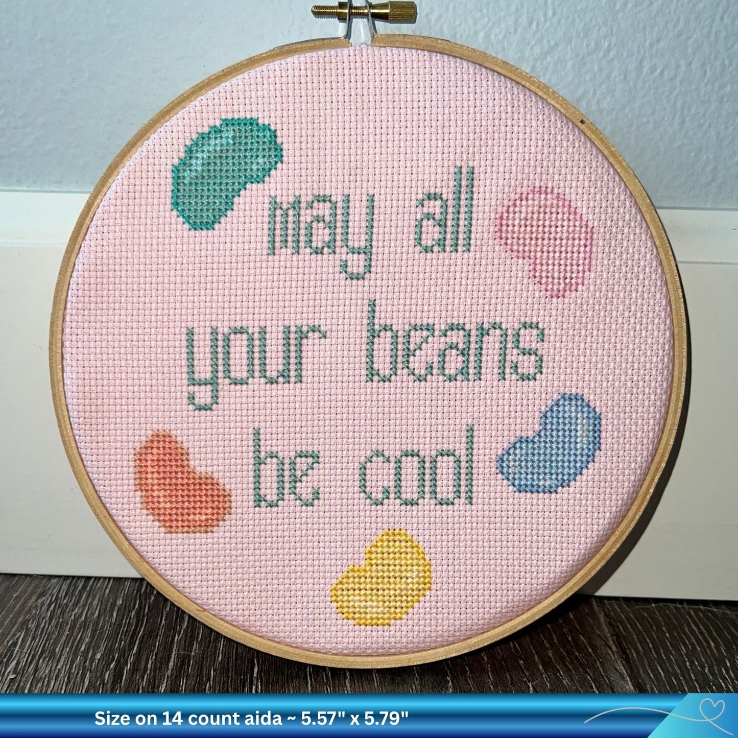 Cross-stitched hoop with 'May all your beans be cool' text on a pink Aida fabric background.