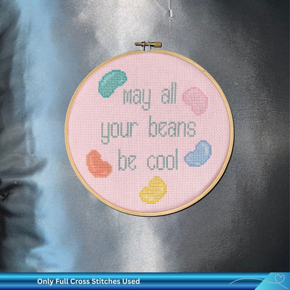 Embroidery hoop with humorous cross-stitched text and colorful beans on a pink Aida fabric against a silver background
