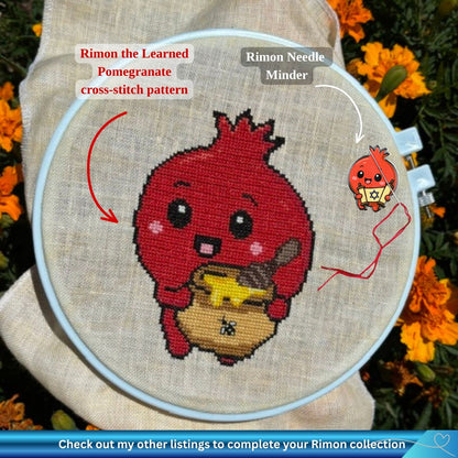 A cross-stitch embroidery hoop holding a finished stitched design of a red pomegranate character (Rimon) holding a honey jar, shown outdoors among orange flowers. Text labels point to the design as 'Rimon the Learned Pomegranate cross-stitch pattern' and to a small matching charm as 'Rimon Needle Minder.