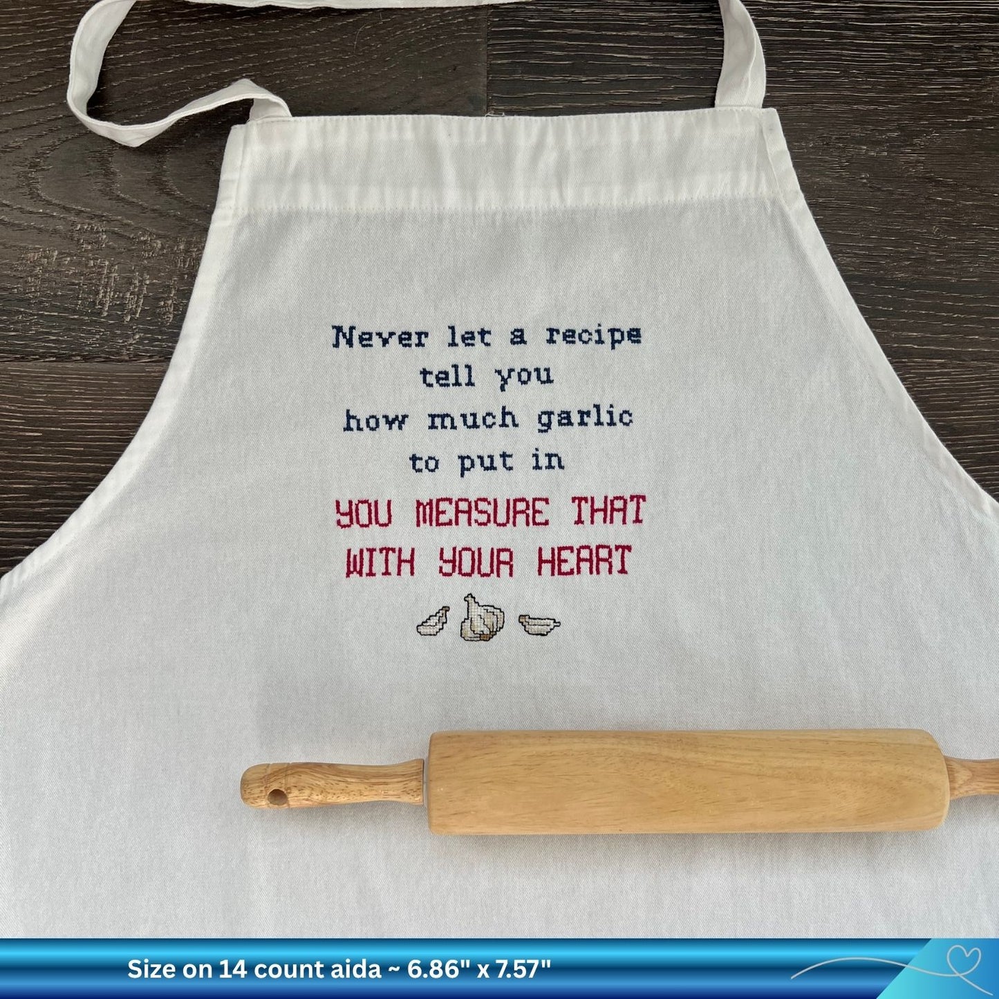 Cross stitched funny saying phrase "never let a recipe tell you how much garlic to put in" stitched onto a white apron. There is also a wooden rolling pin resting on the white apron.
