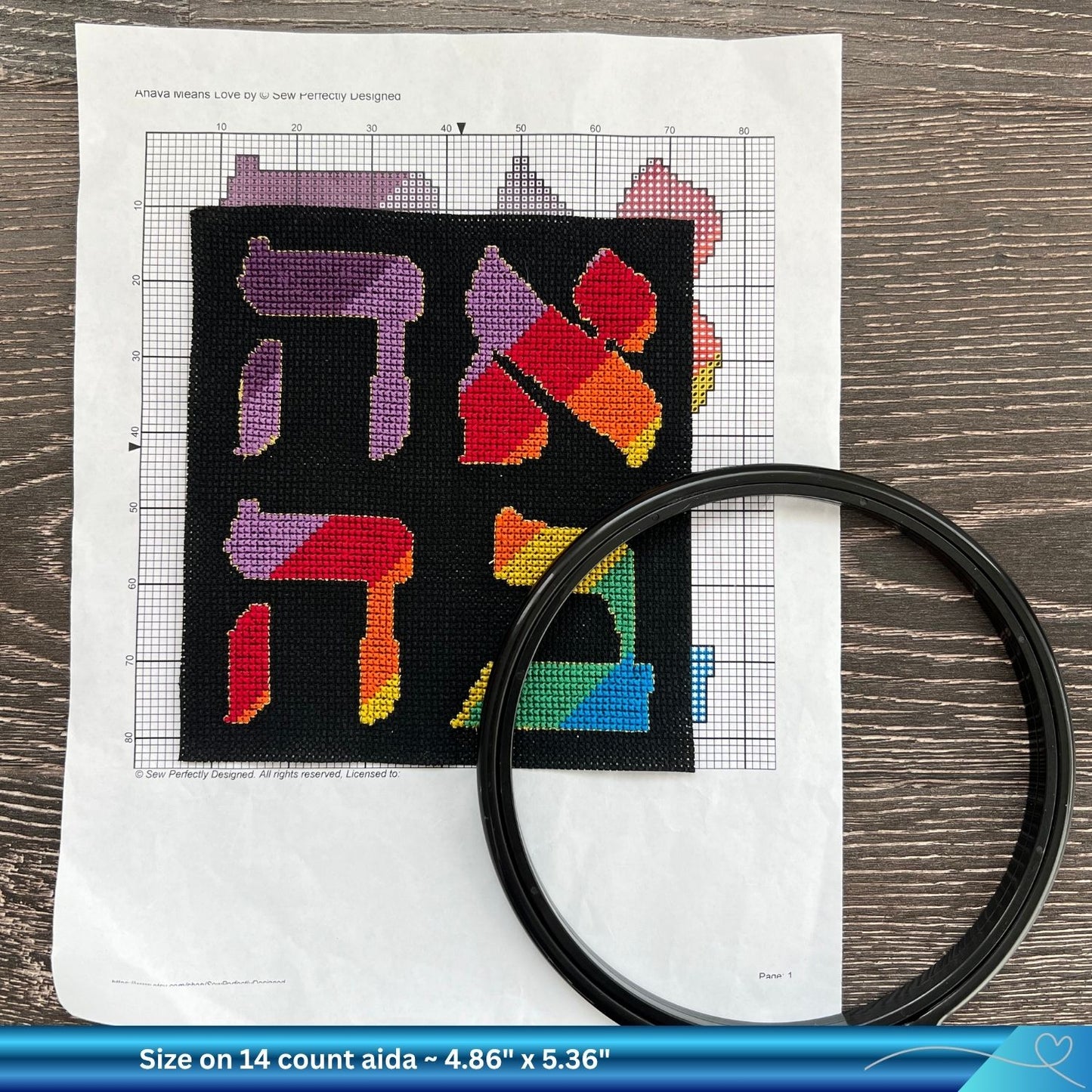 Cross-stitched word Ahava in Hebrew against a print out of a pattern and a black embroidery hoop