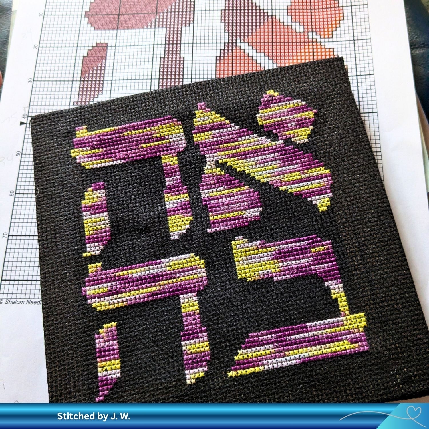Cross-stitched design of the Hebrew word for Love (Ahava) with colorful letters Hebrew on black fabric, placed on a printed out pattern.