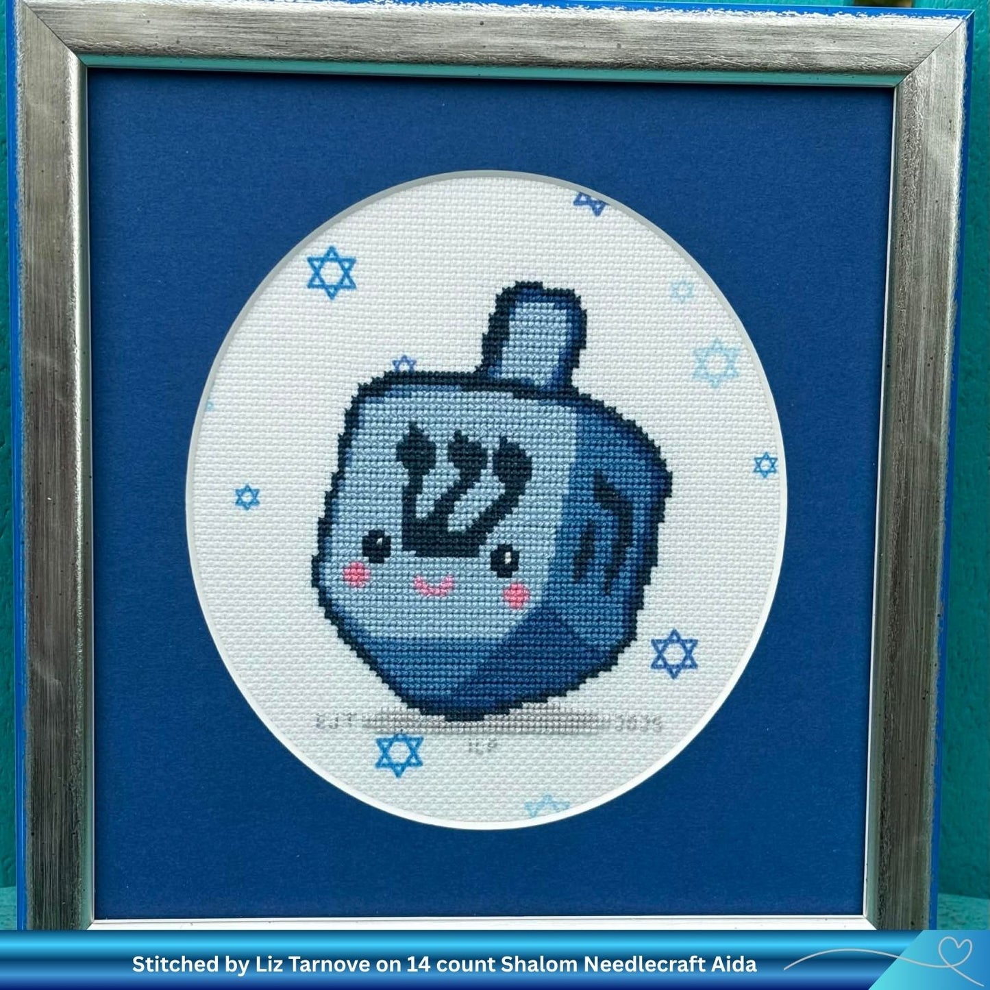 Cross-stitched design of Shlomo the Hanukkah dreidel framed with a blue border