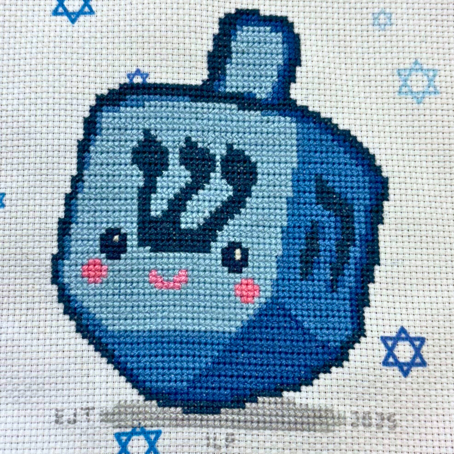 Cross-stitched blue dreidel with a face on a white background with blue star patterns