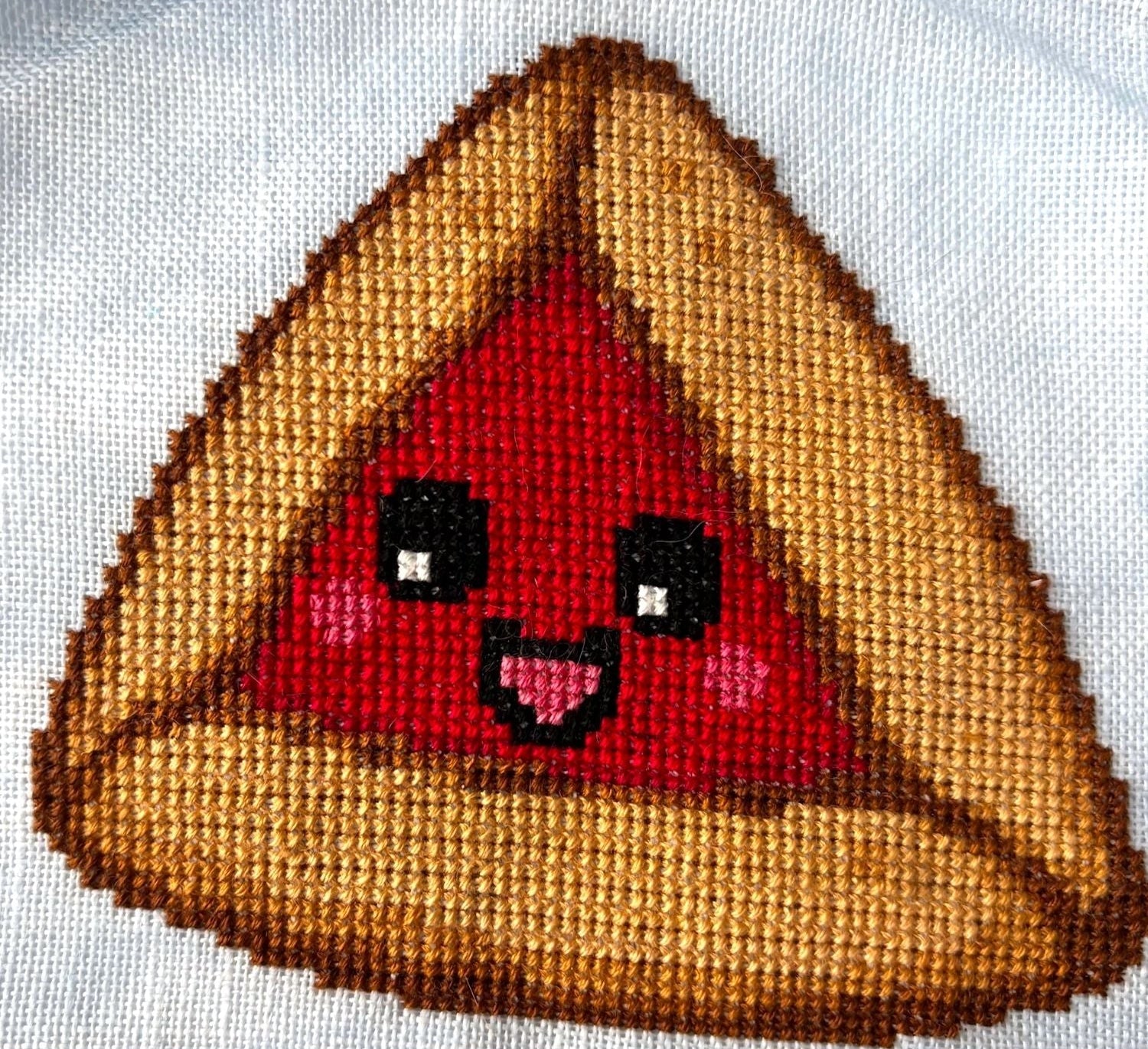 Cross-stitched design of a smiling Hamantaschen character on brown fabric.