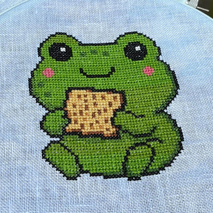 Cross-stitched frog holding a Matzos cracker on light blue fabric.