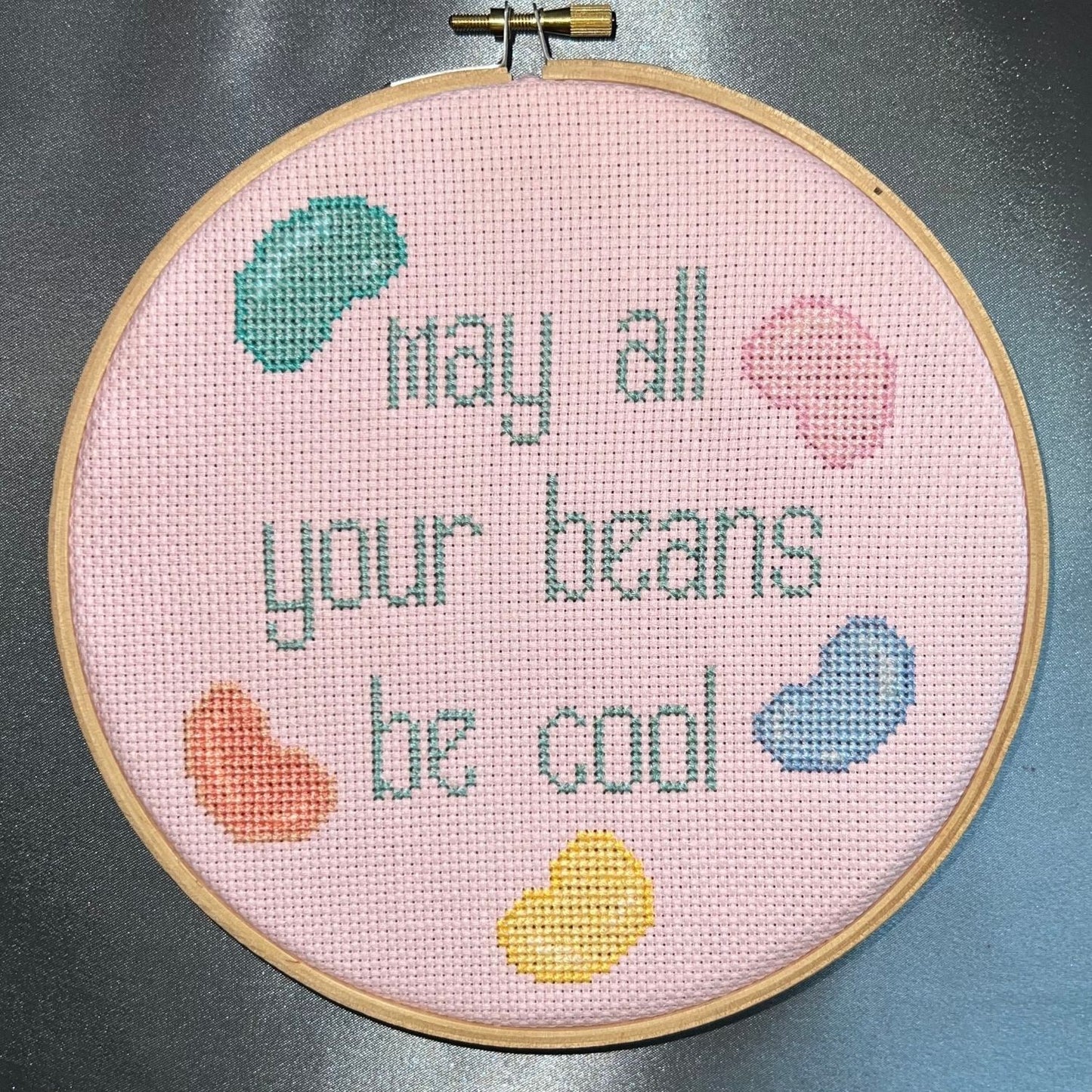 Snarky cross stitch design saying "may all your beans be cool" with jelly beans surrounding it frame in a wooden embroidery hoop