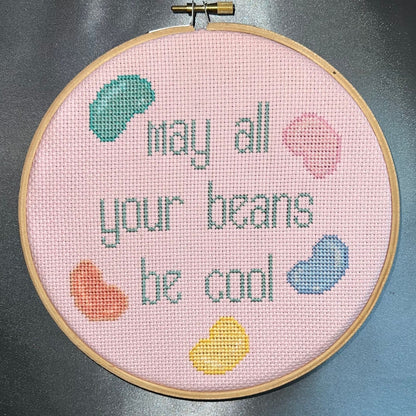 Snarky cross stitch design saying "may all your beans be cool" with jelly beans surrounding it frame in a wooden embroidery hoop