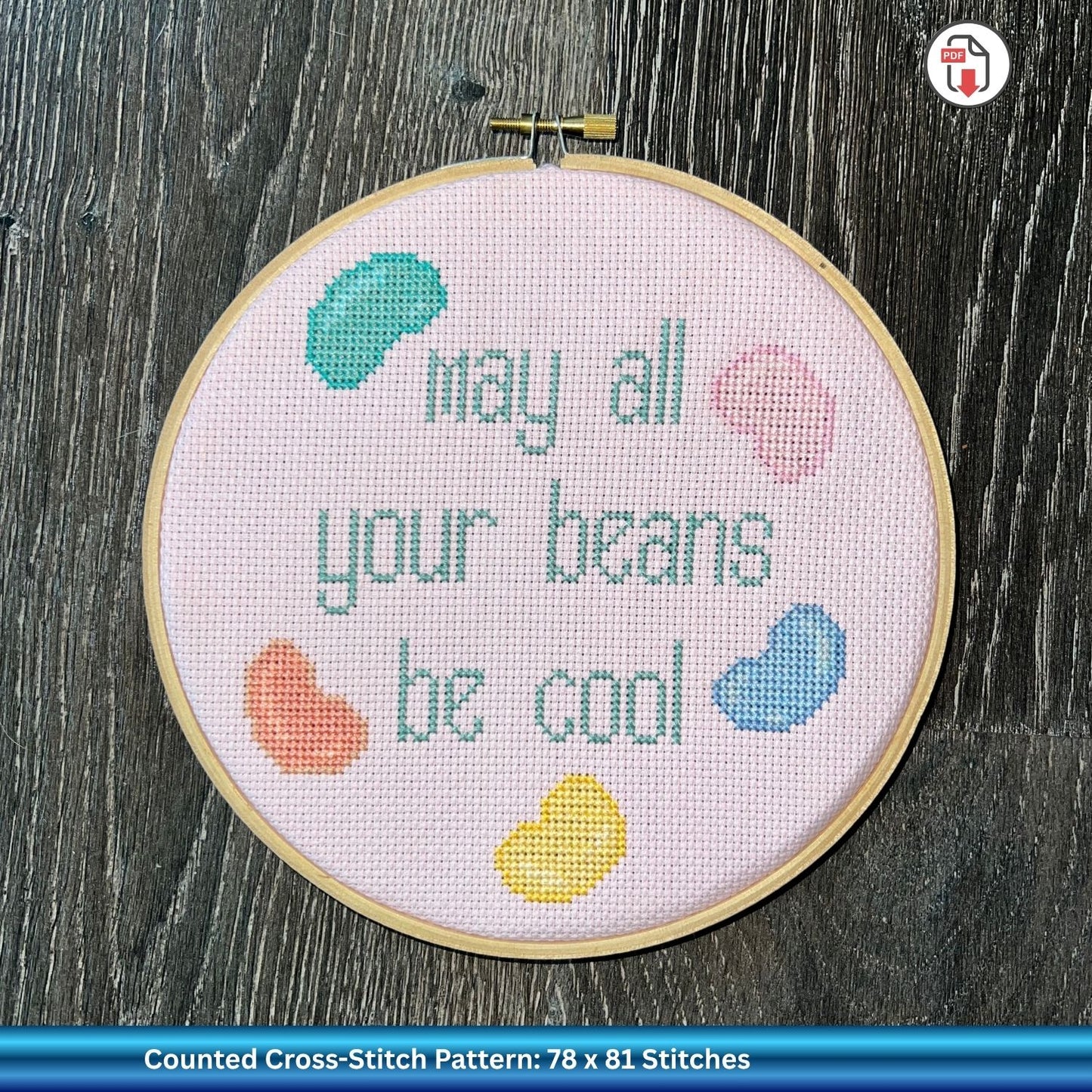 Cross-stitched hoop with 'May all your beans be cool' text and colorful beans on a pink background.