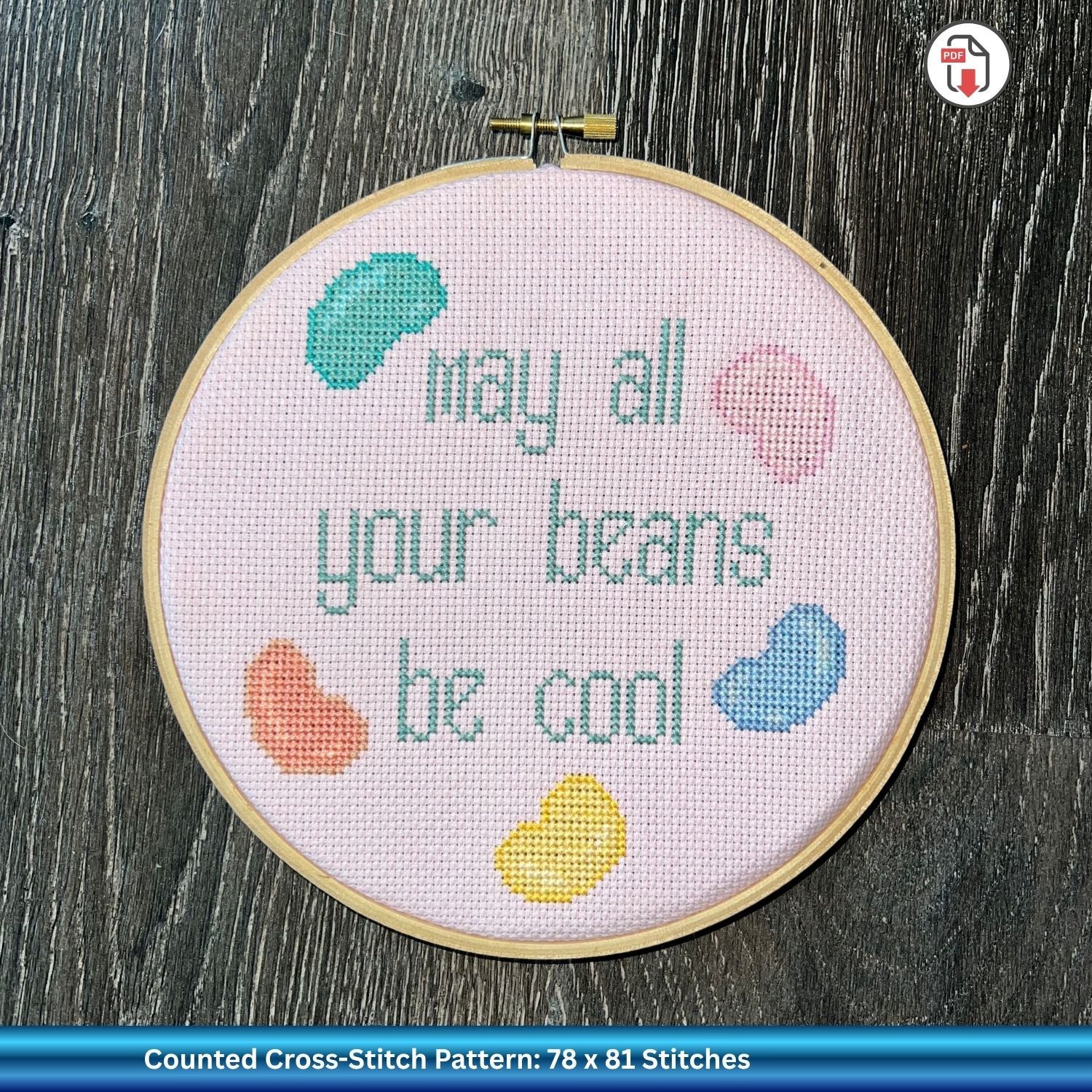 Cross-stitched hoop with 'May all your beans be cool' text and colorful beans on a pink background.