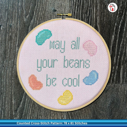 Cross-stitched hoop with 'May all your beans be cool' text and colorful beans on a pink background.