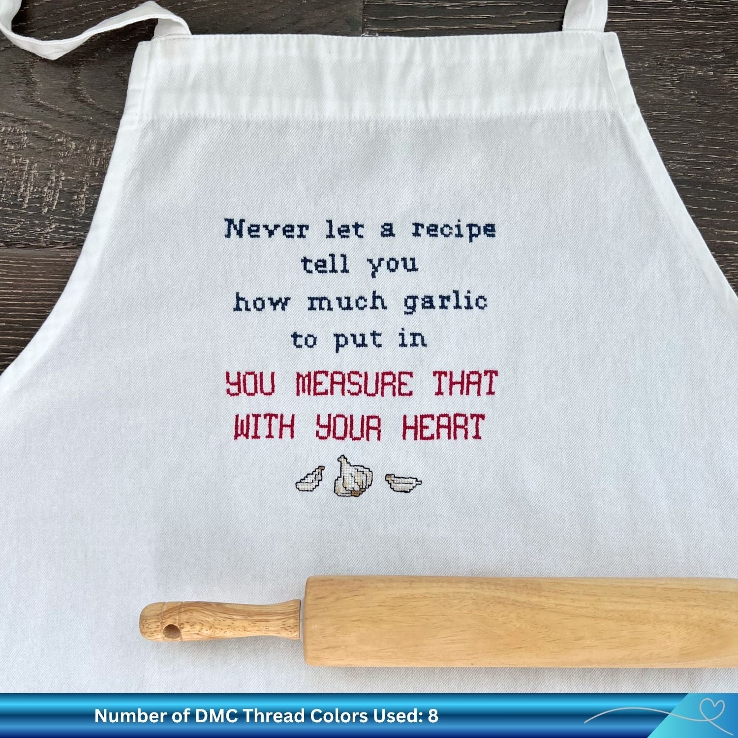 White apron with the phrase "never let a recipe tell you how much garlic to put in" stitched onto the front. There is also a wooden rolling pin resting on the white apron.