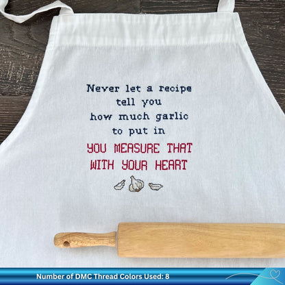 White apron with the phrase "never let a recipe tell you how much garlic to put in" stitched onto the front. There is also a wooden rolling pin resting on the white apron.