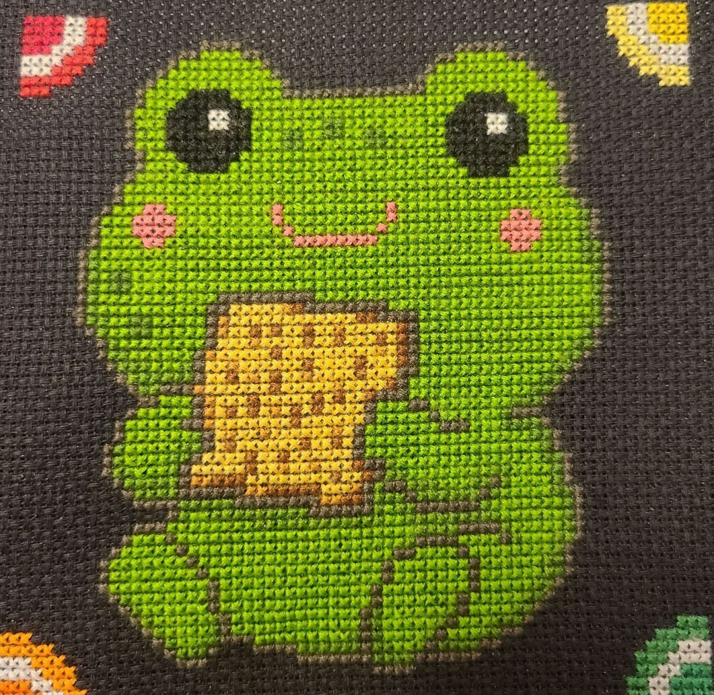 Cross-stitched green frog on black fabric with text indicating stitching details.