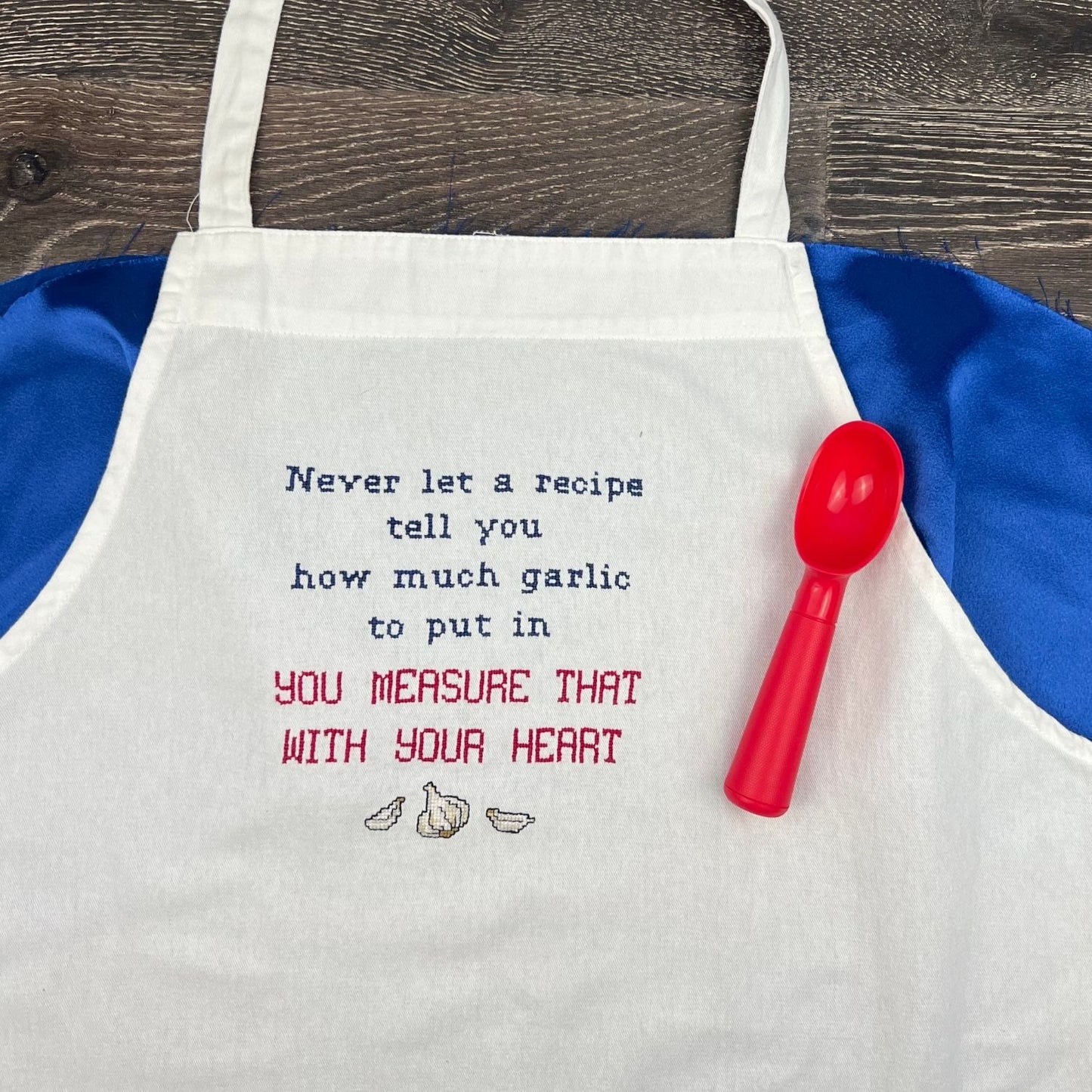 Hand embroidered apron with the phrase "never let a recipe tell you how much garlic to put in" cross stitched onto the front. There is also a red scoop next to the white apron.