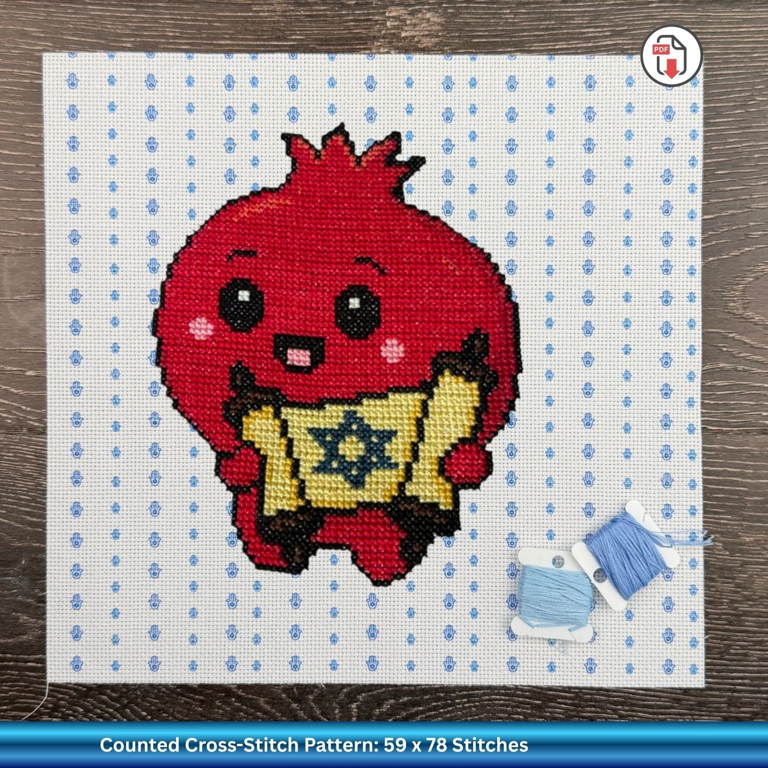 Cross-stitched pomegranate character holding a Torah on Aida fabric with blue Hamsas printed