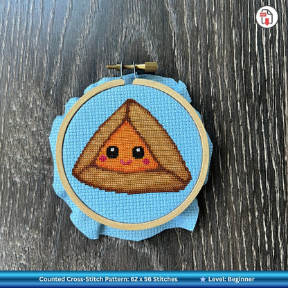Cross-stitched design of a smiling orange hamantaschen character in an embroidery hoop